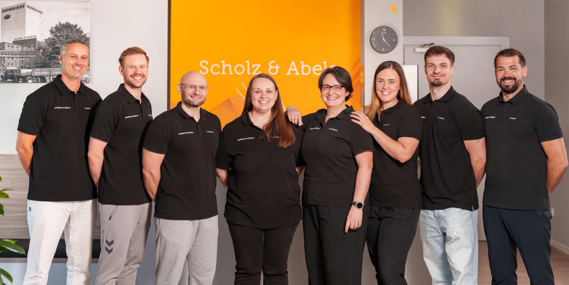 Szenenbild - Team der Physiotherapiepraxis Scholz & Abela stehend Gruppenbild des Scholz & Abela Team, stehend nebeneinander.